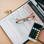 Tampilan dari atas meja kerja dengan dokumen rumus harga, kalkulator, dan kacamata sebagai ilustrasi perhitungan biaya total rata-rata